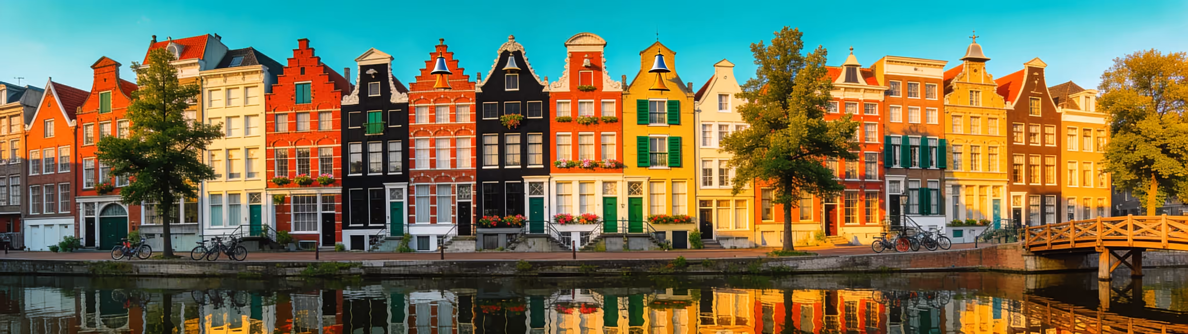 A long row of tall, narrow canal houses with step and bell gables mirrored in the still water of an Amsterdam canal, a small wooden bridge at the end